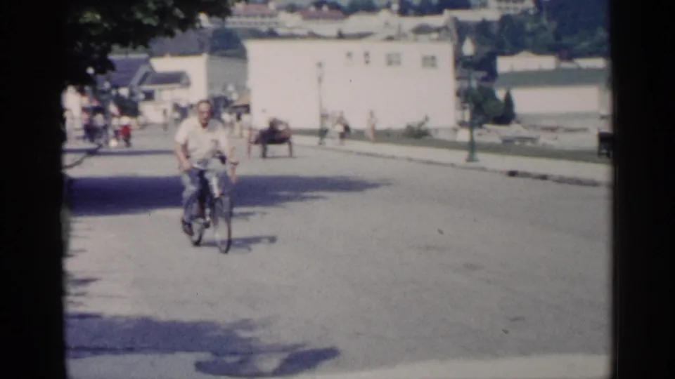 An adult man rides a bicycle down a quiet Michigan waterfront street, with piers, buildings, and pedestrians in the background.