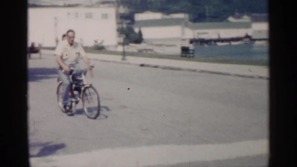 A man in a light shirt and glasses rides a bicycle on a paved street near the Michigan lakeshore and waterfront structures.