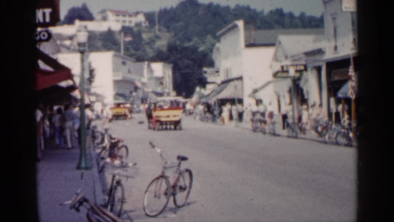 Small-town street with pedestrians, bicycles, storefronts, and a yellow vehicle.