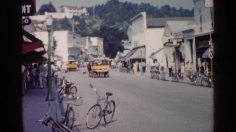 1960 Michigan 8mm frame showing a busy small-town street with storefronts, pedestrians, curbside bicycles, and a yellow vehicle in the road.