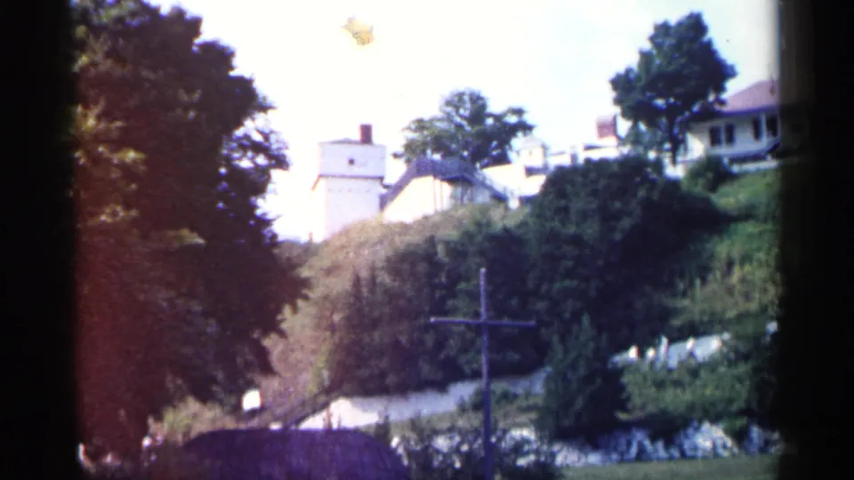 1960 Michigan 8mm frame of hillside buildings partly obscured by trees, with a wooden cross and dark roofline in the lower foreground.