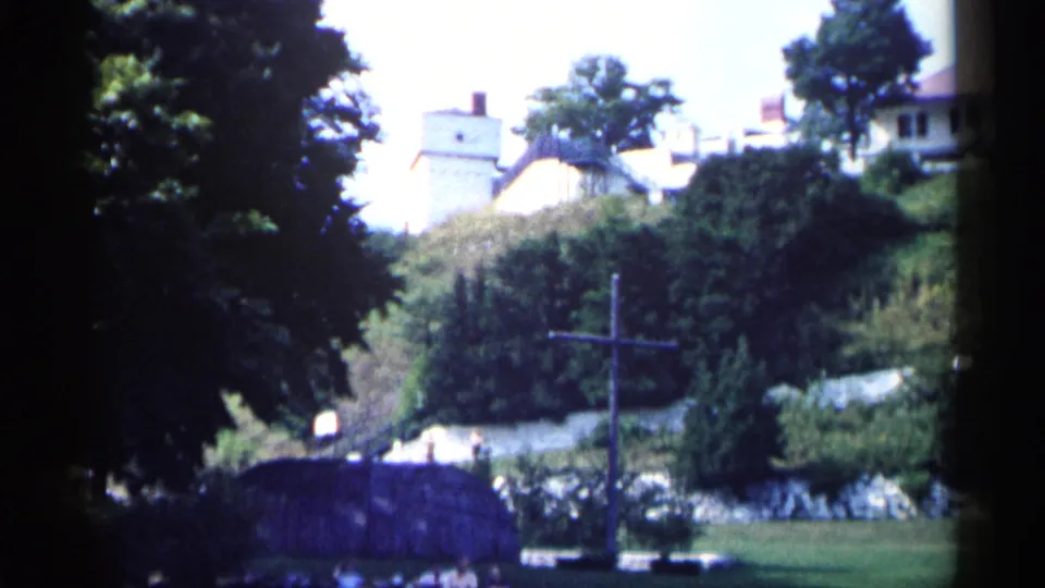 1960 Michigan 8mm frame showing pale buildings on a green hillside, a foreground cross, trees, and a dark roofline at the bottom edge.