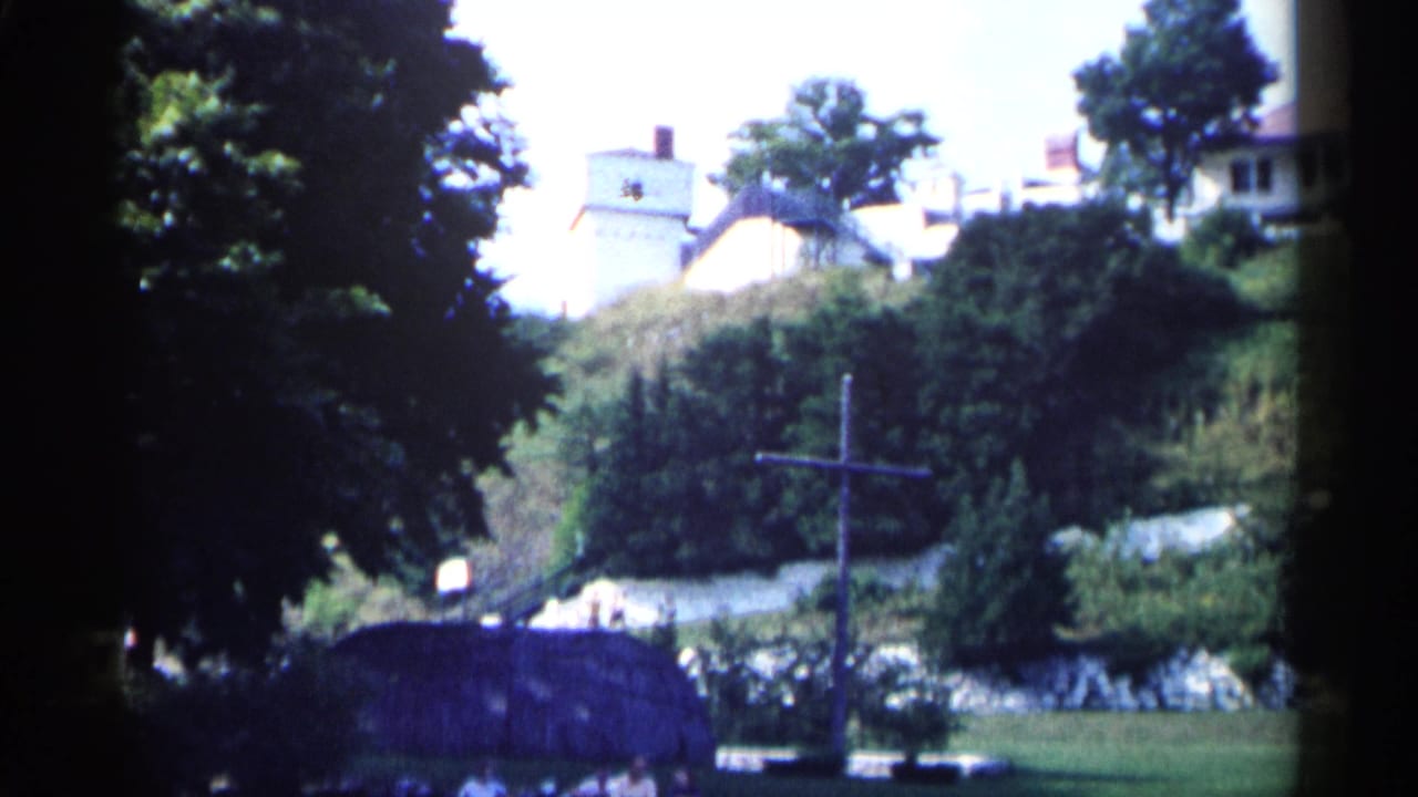 Wide view of trees, a hillside, distant houses, and a wooden cross near the foreground.