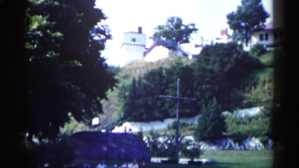 1960 Michigan 8mm frame showing leafy trees, a sloping landscaped area, distant light houses, and a dark wooden cross in the foreground.
