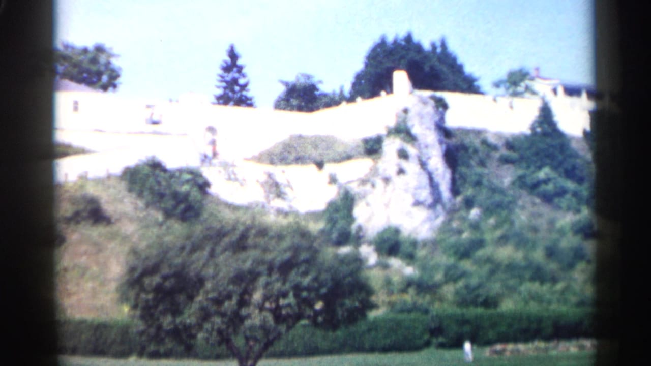 White hillside wall, rocky outcrop, shrubs, trees, and small distant figures.