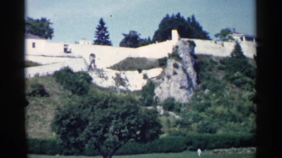 1960 Michigan 8mm frame showing a white structure on a green hillside with an arched detail, rocky slope, hedges, and trees.