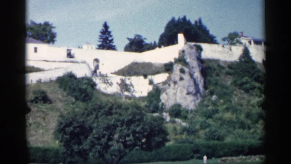 1960 Michigan home-movie frame of a white hillside structure, stone outcrop, green landscaping, trees, and pale sky.
