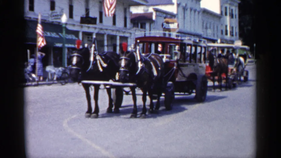 1960 Michigan street scene with two dark horses pulling an open carriage, storefronts, American flags, pedestrians, and another carriage behind.