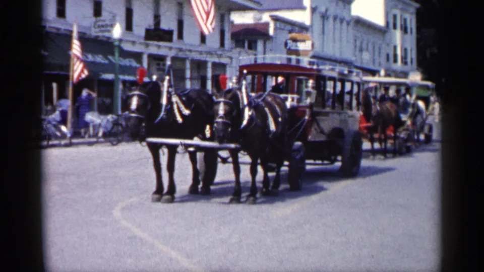 1960 Michigan home-movie frame of a horse team and open carriage on a paved downtown street with flags, storefront signs, and pedestrians.