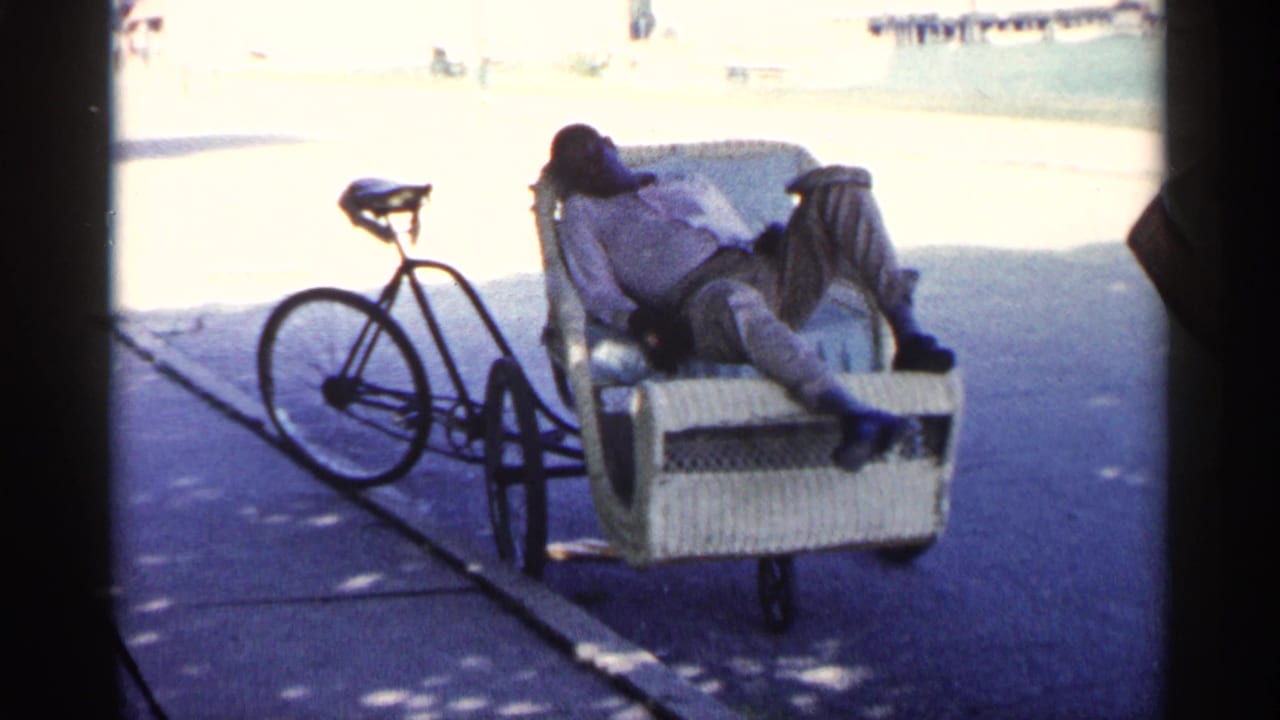 Man reclines asleep in a wicker bicycle passenger cart beside a parked bicycle on pavement.