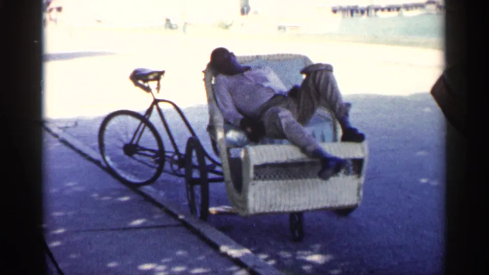 In 1960 Michigan home-movie footage, a man appears to sleep in a wicker bicycle cart parked beside a bicycle on a sunlit paved area.