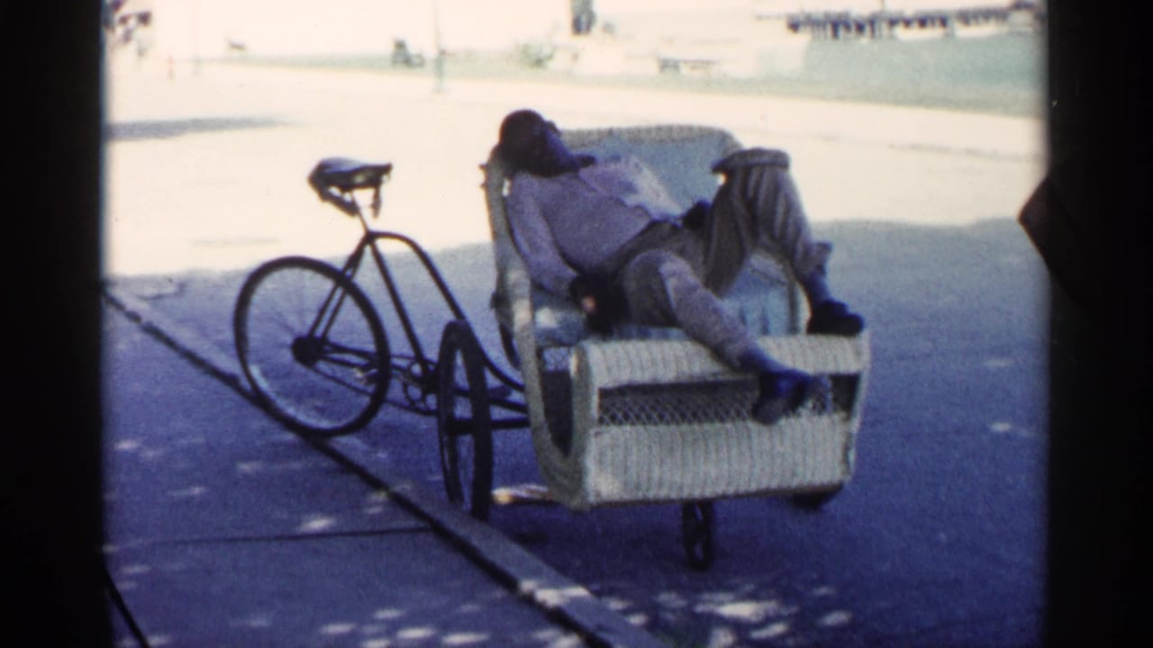 The home-movie frame shows a man reclining in a wicker bicycle cart, the bicycle parked alongside on a bright paved surface.