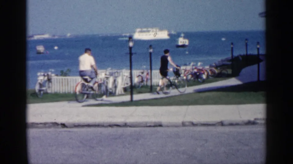 In the Michigan waterfront scene, two adults ride bicycles along a path while boats and a ferry-like vessel appear on the water beyond a railing.