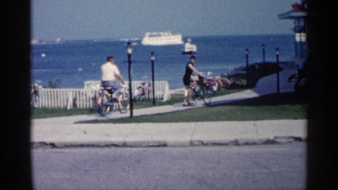 1960 Michigan lakeside home-movie frame showing two cyclists near a picket fence, lampposts, blue water, and a distant white ferry.