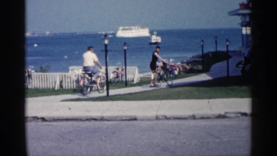 1960 Michigan lakeside home-movie frame showing two cyclists near a picket fence, lampposts, blue water, and a distant white ferry.
