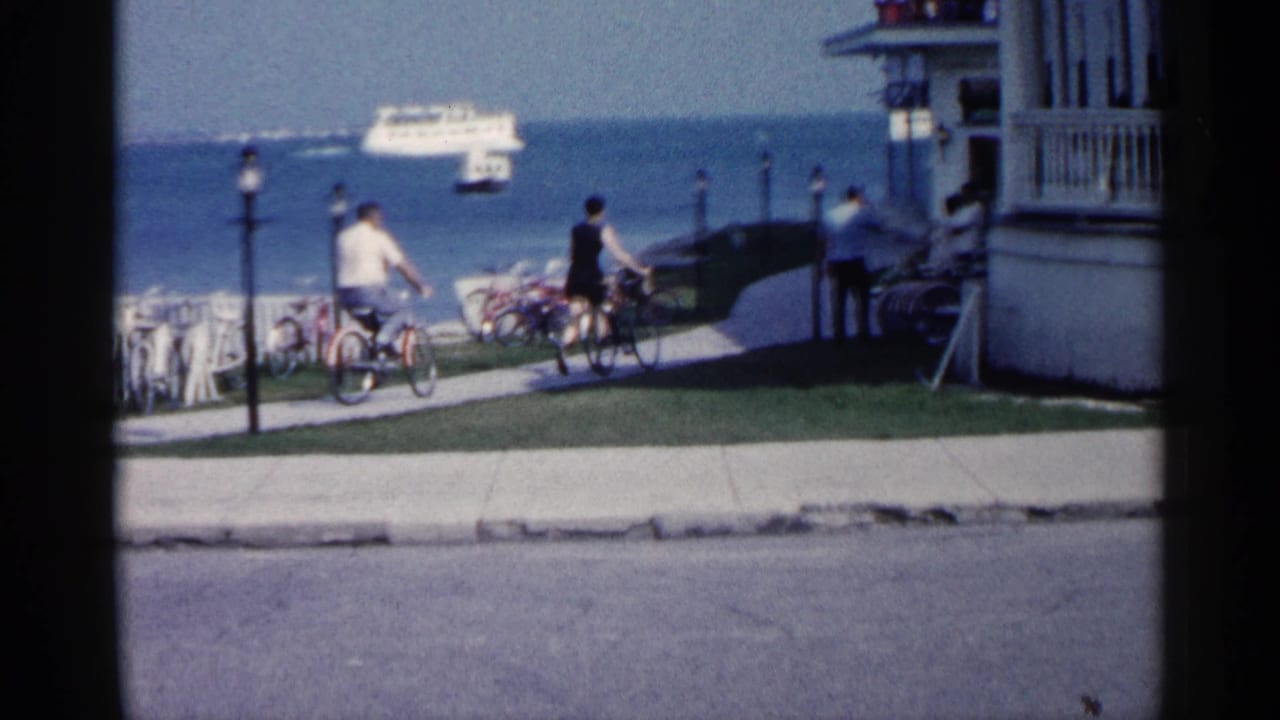 Cyclists continue beside the lake near a porch-front building and parked bicycles.