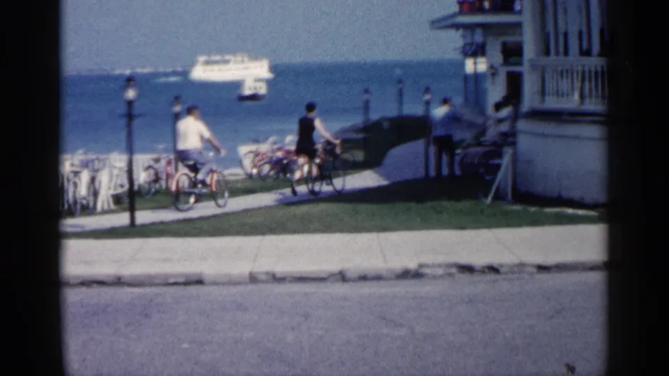 1960 Michigan waterfront frame with cyclists on a paved path, parked bicycles, lampposts, a porch-front building, and a ferry in the distance.