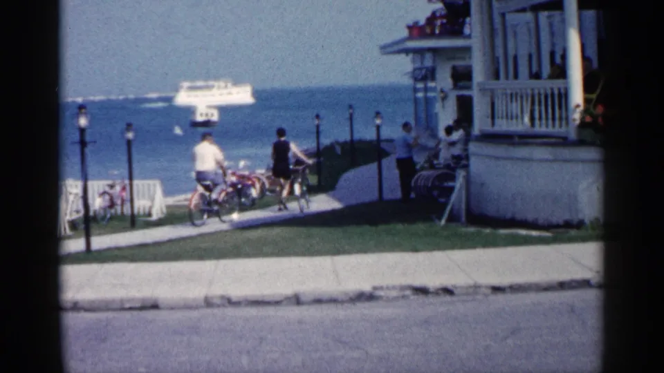 1960 Michigan lakeside view with bicyclists on a path, a white picket fence, porch columns, seated people, blue water, and a distant ferry.