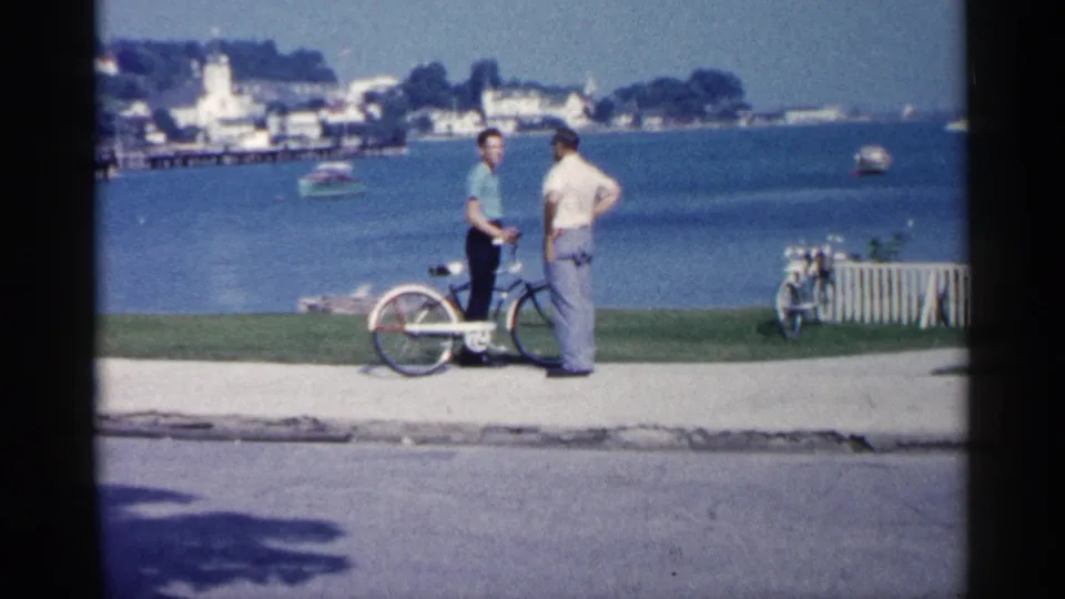 1960 Michigan lakeside frame showing two men by a bicycle on a paved path, with open water, distant buildings, and a shoreline behind them.