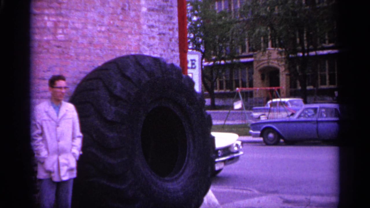In a Michigan 1960s street scene, a man with glasses stands next to a huge truck tire with period cars behind him.