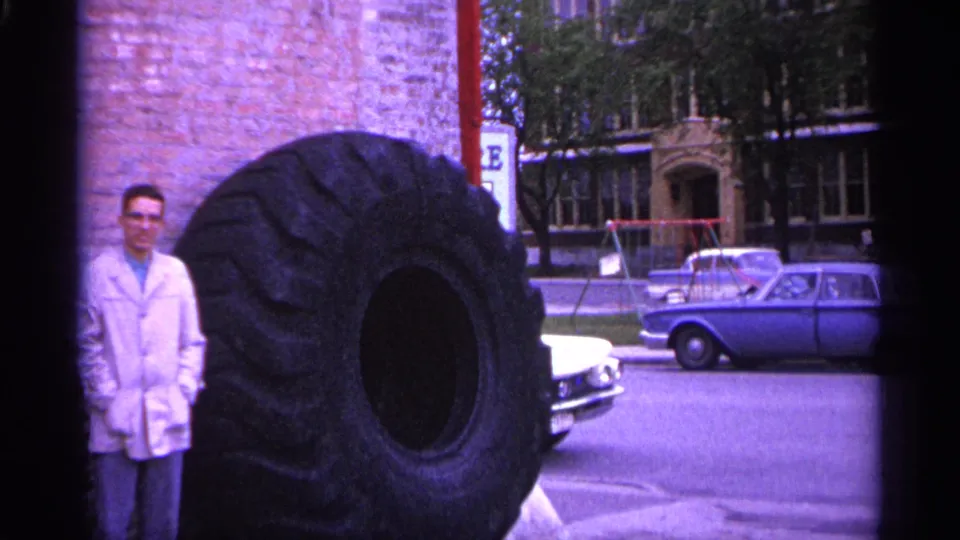 The man stands near the edge of a very large truck tire, showing the scale difference in a 1960s Michigan street view.