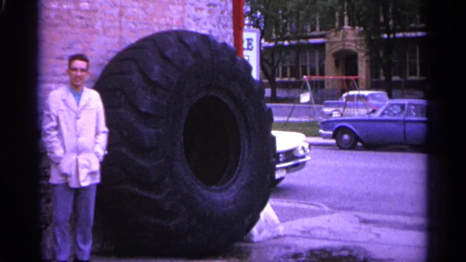 A man poses beside a massive truck tire in a 1960s Michigan street scene, with classic cars and masonry buildings behind him.