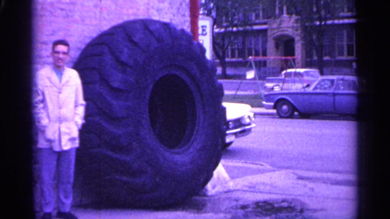 Man in a white coat stands beside a massive industrial tire on a city street.