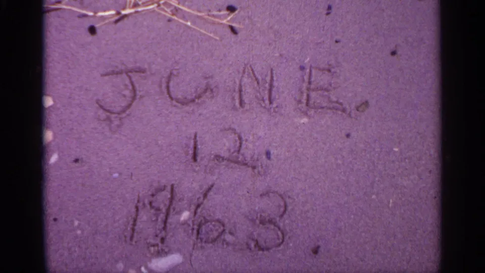 Close view of sand marked with a handwritten date, with dry twigs and visible 8mm color shift.