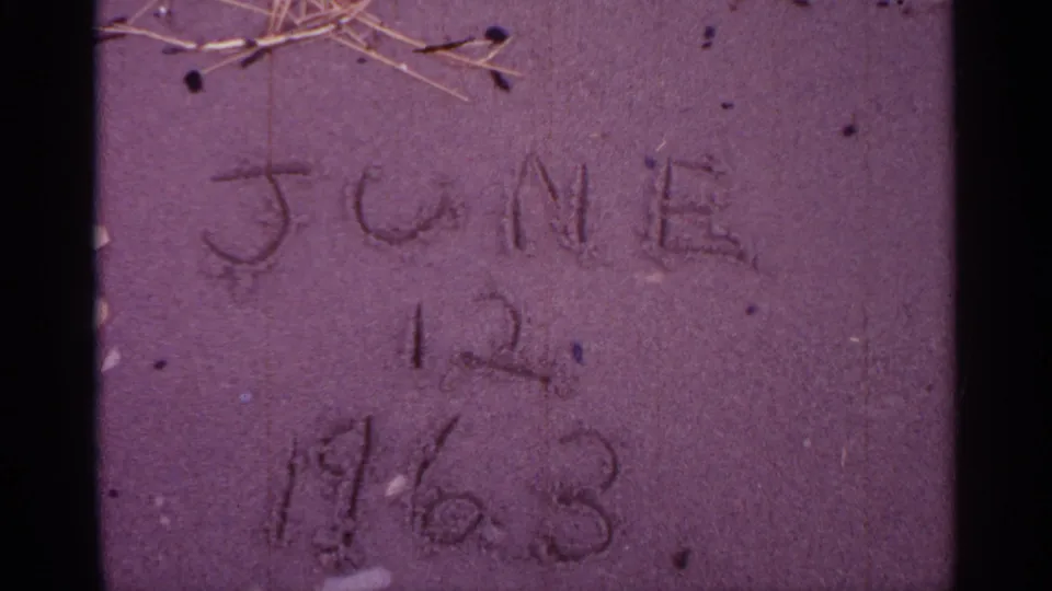 Sand close-up showing the scratched date more centrally, with twigs above and visible 8mm grain.