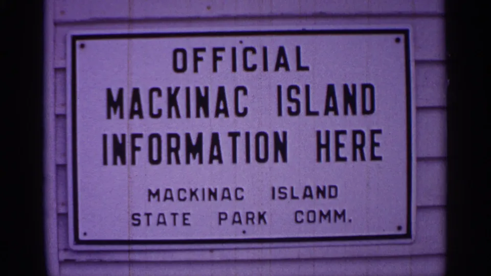 1960 Michigan 8mm frame showing an official Mackinac Island information sign on a light building facade with dark film edges.
