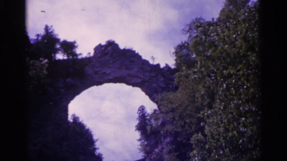 1960 Michigan 8mm view of a natural rock arch surrounded by leafy trees under a cloudy sky on Mackinac Island.