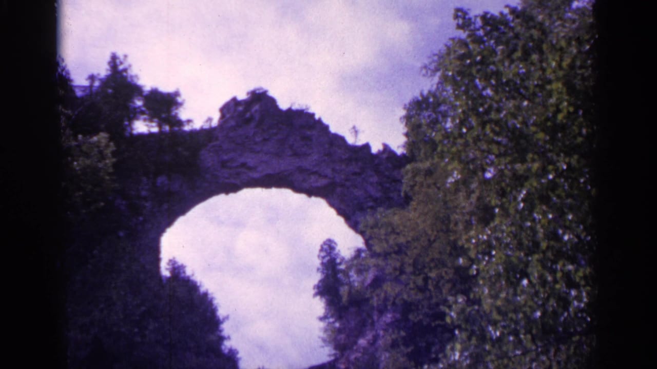 Rocky arch or bridge-like formation above trees with cloudy sky behind it.