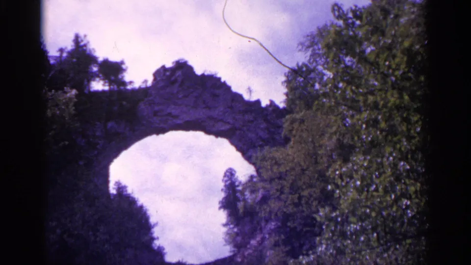 1960 Michigan home-movie frame with a rocky arch or bridge-like formation, surrounding trees, and visible film gate edges.