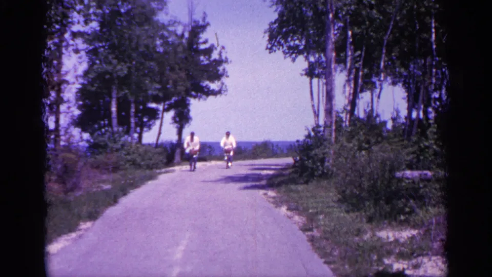 1960 Michigan 8mm frame showing two cyclists on a quiet paved wooded path with distant water visible ahead.