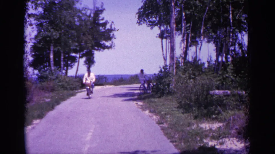 A cyclist continues forward while the second rider turns near the road edge in 1960 Michigan home-movie footage.