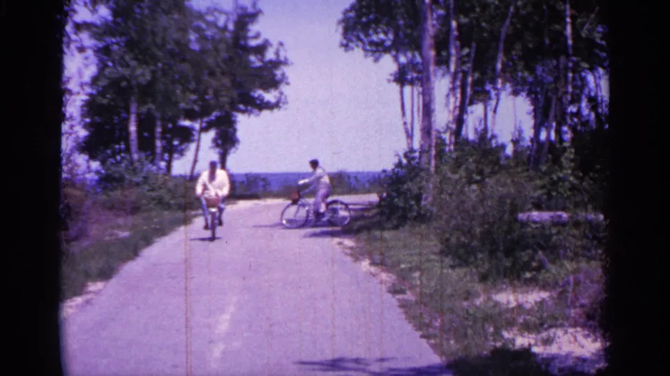 One bicyclist rides toward the camera as another crosses sideways on the wooded Michigan road, with visible 8mm grain and color cast.
