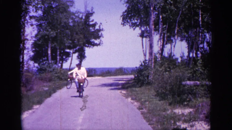 1960 Michigan frame with an adult cyclist riding toward the camera and a second cyclist turning farther back on the wooded path.