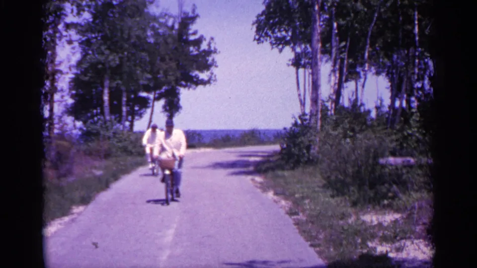 1960 Michigan home-movie frame of two adults bicycling toward the camera on a paved path lined with trees and low vegetation.