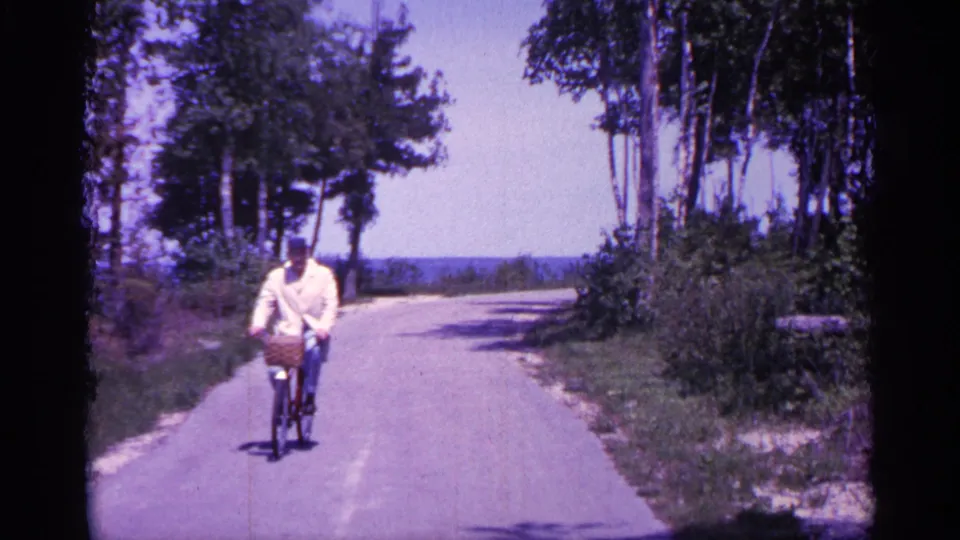 1960 Michigan frame showing an adult cyclist with a front basket riding close to the camera on a sunlit wooded path.