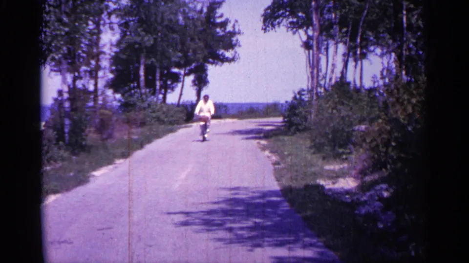 The cyclist, wearing a light-colored shirt, approaches on an empty Michigan road with trees and water visible in the distance.