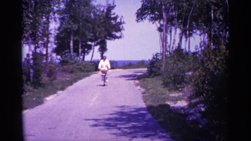 A cyclist continues toward the camera on a sunlit paved road, with dark tree shadows falling across the foreground.