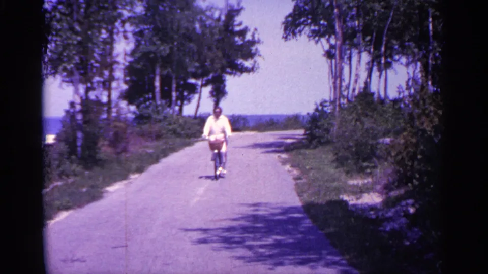 Near the end of the 1960 Michigan clip, the cyclist is closer to camera on the tree-lined road with water beyond the bend.