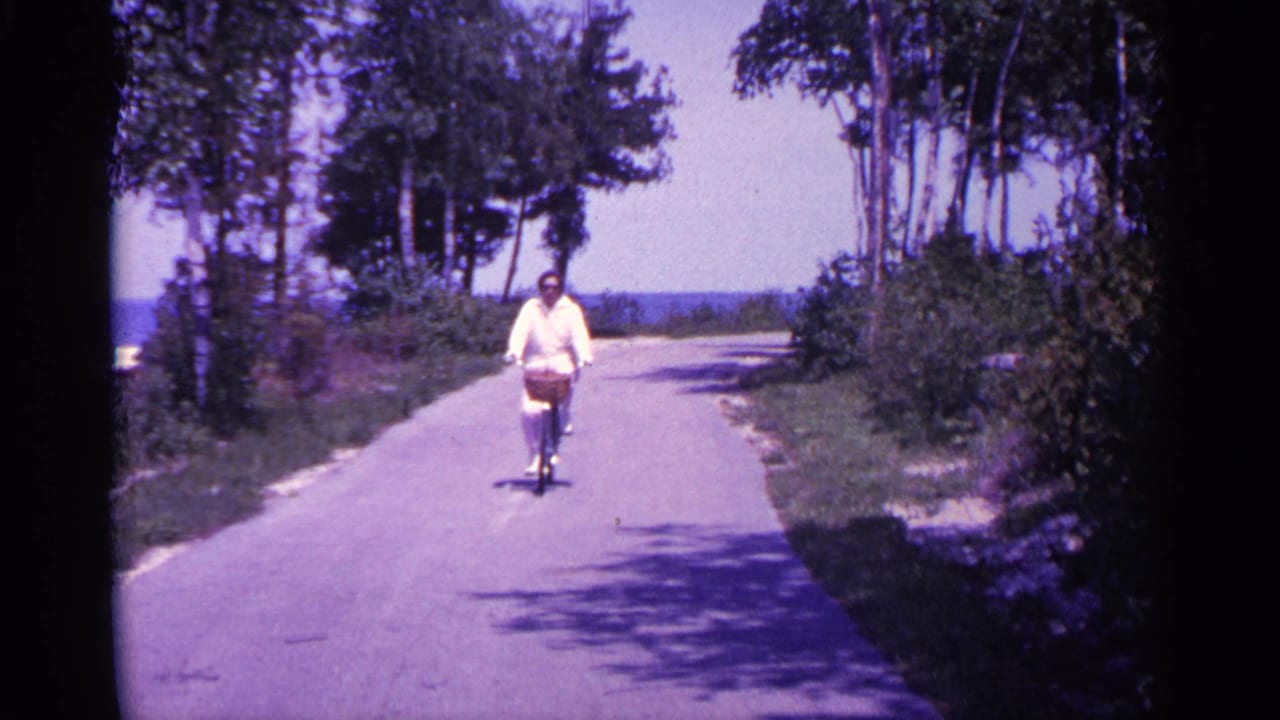 Woman rides a bicycle on a paved tree-lined path with water visible in the distance.