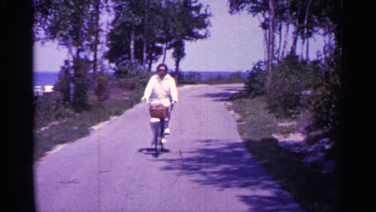 Woman in a white jacket pedals closer on a bicycle with a basket along the path.