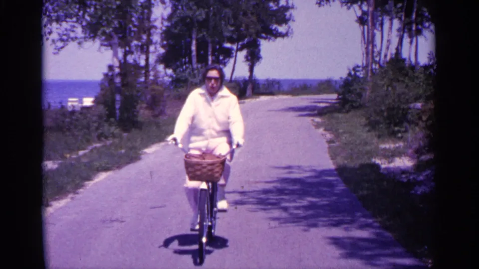 A closer frame shows the adult rider in sunglasses and a white jacket steering a bicycle with a front basket.