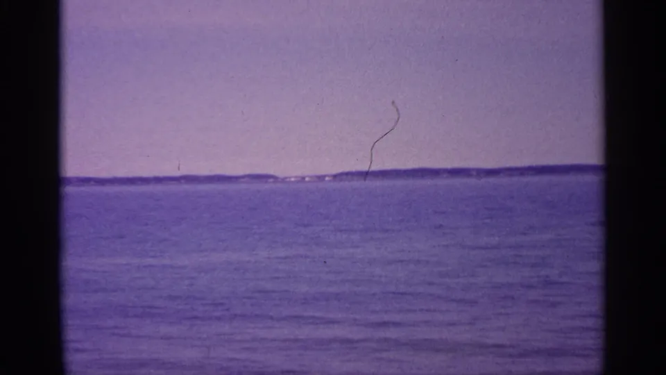 1960 Michigan home-movie frame with open water, distant shoreline, faint buildings, and visible film wear.