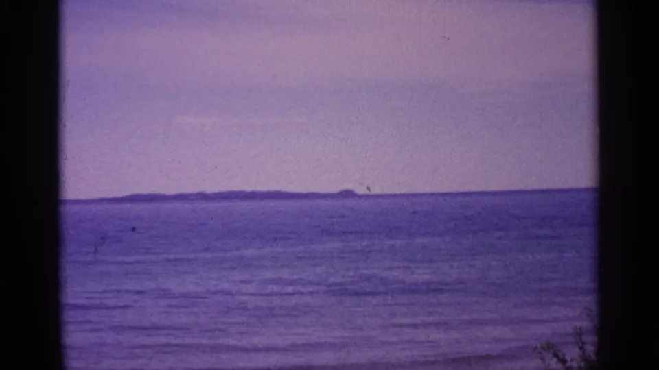 1960 Michigan home-movie frame looking over calm water toward a low distant landmass, with faint vegetation at the lower edge.