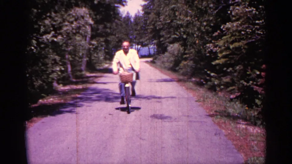 An older man in light clothing rides closer to the camera, his bicycle basket visible against the sunlit road and leafy roadside.