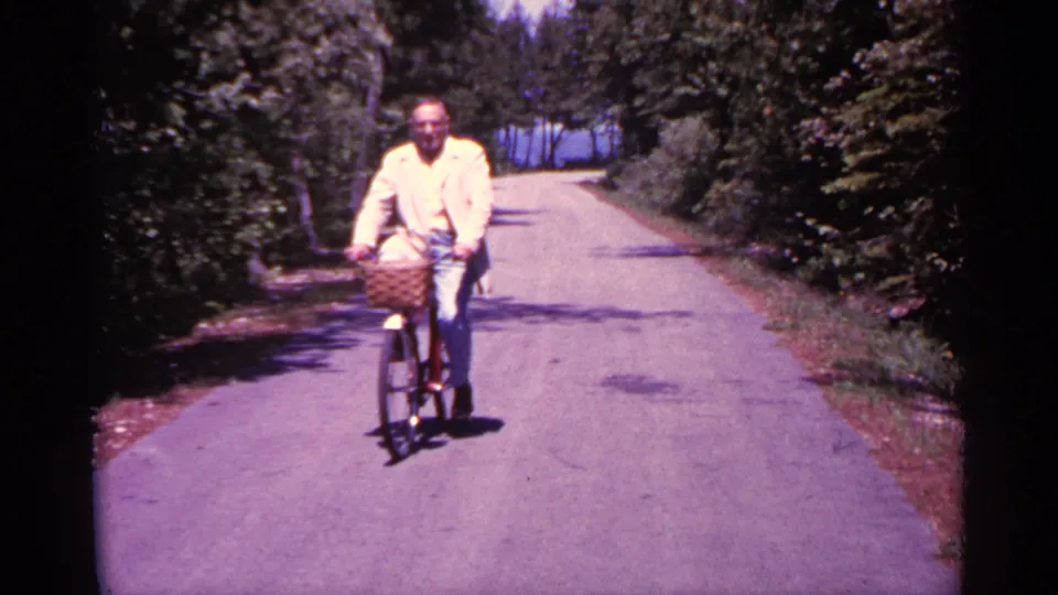 Near the end of the shot, the man rides close to the camera on the Michigan road, framed by trees and strong midday light.