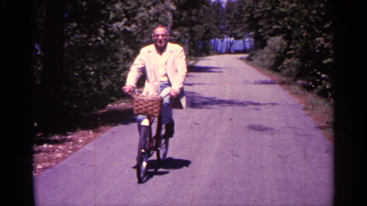 Man in a light jacket rides a bicycle toward the camera on a tree-lined road.